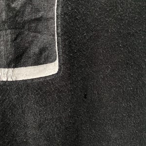 May include: Close-up of a black garment with a rectangular patch and a white trim. The fabric has a textured appearance. The patch is a darker shade of black and has a reflective quality.
