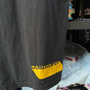 Pu&ograve; includere: Maglietta nera con logo "QUIKSILVER" giallo in basso a destra.