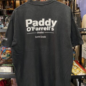 May include: Black t-shirt with white text that reads "Paddy O'Farrell's Beaches Toronto Canada". The shirt is hanging on a wooden hanger. The shirt appears to be vintage.