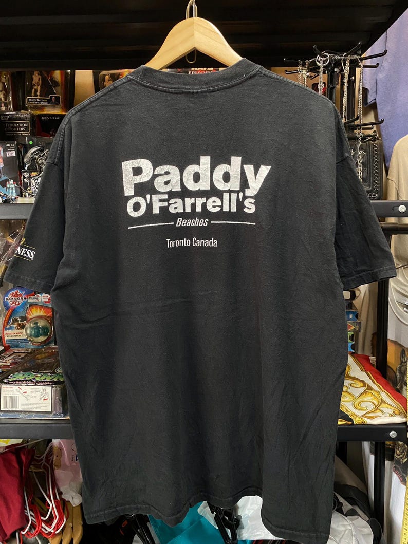 May include: Black t-shirt with white text that reads "Paddy O'Farrell's Beaches Toronto Canada". The shirt is hanging on a wooden hanger. The shirt appears to be vintage.