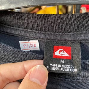 Pu&ograve; includere: Maglietta nera con un'etichetta bianca che dice "21/28" e un'etichetta nera con un logo rosso e il testo "M MADE IN MEXICO/FABRIQUE AU MEXIQUE"