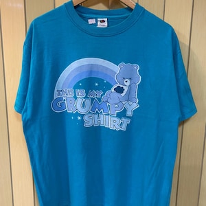 May include: Teal-colored t-shirt with a graphic featuring a grumpy bear character, a rainbow, and the text "THIS IS MY GRUMPY SHIRT." The shirt is hanging on a wooden hanger.