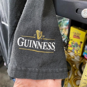 May include: Dark gray t-shirt sleeve featuring the Guinness logo in white and gold. The logo includes a harp symbol and the text "GUINNESS" with the years "1759". The sleeve is made of a soft, textured fabric.