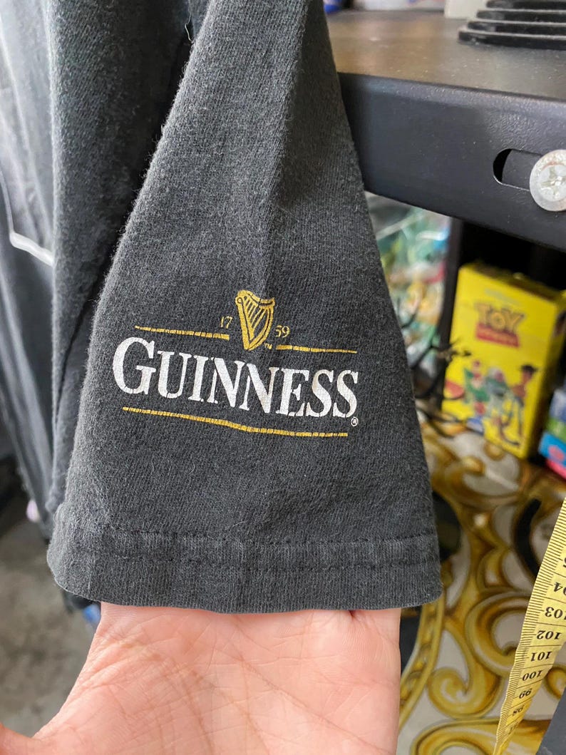 May include: Dark gray t-shirt sleeve featuring the Guinness logo in white and gold. The logo includes a harp symbol and the text "GUINNESS" with the years "1759". The sleeve is made of a soft, textured fabric.