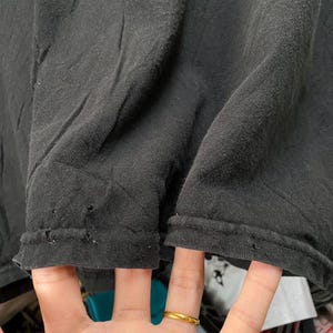 May include: Close-up of a black t-shirt, showing the hem and the fabric's texture. The fabric is slightly wrinkled, with small holes near the hem. A hand with a gold ring is visible.