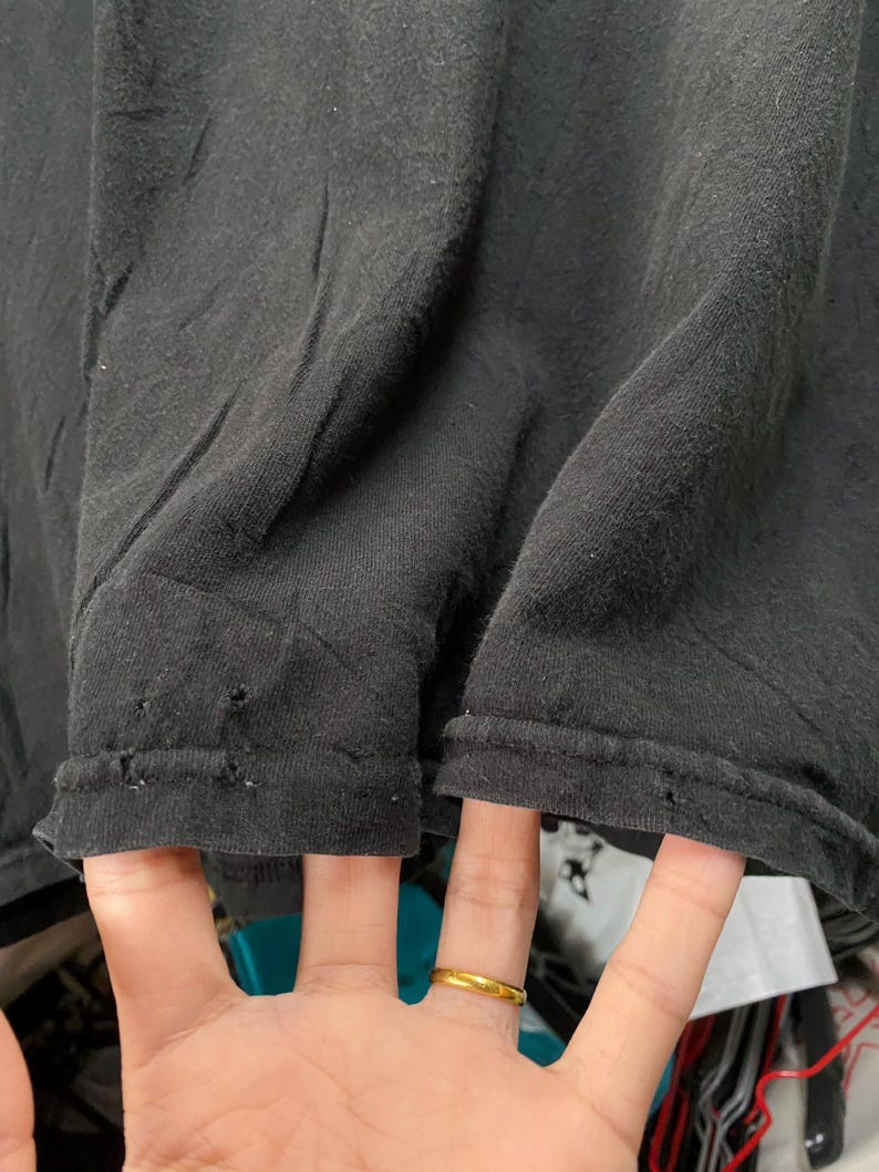 May include: Close-up of a black t-shirt, showing the hem and the fabric's texture. The fabric is slightly wrinkled, with small holes near the hem. A hand with a gold ring is visible.