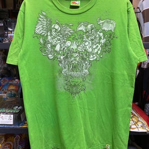 Può includere: T-shirt verde brillante con un design grafico dettagliato nelle tonalità del verde. Il design include figure di teschi e angeli con dettagli intricati. La maglietta ha un girocollo e maniche corte.