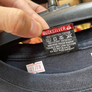 Pu&ograve; includere: Maglietta nera con un'etichetta bianca che dice "Quiksilver" e contiene istruzioni per la cura. L'etichetta ha anche un piccolo logo di un'onda. La maglietta ha un'etichetta bianca con il numero "21/28" su di essa.