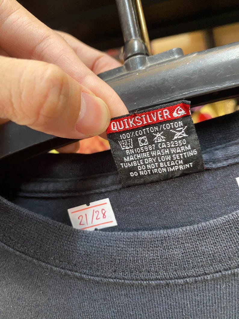 Pu&ograve; includere: Maglietta nera con un'etichetta bianca che dice "Quiksilver" e contiene istruzioni per la cura. L'etichetta ha anche un piccolo logo di un'onda. La maglietta ha un'etichetta bianca con il numero "21/28" su di essa.