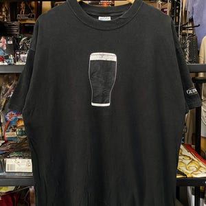 May include: Black t-shirt featuring a white outline of a pint of stout. The shirt has a crew neck and short sleeves. The word "GUIN" is printed on the right sleeve. The shirt is hanging on a wooden hanger.