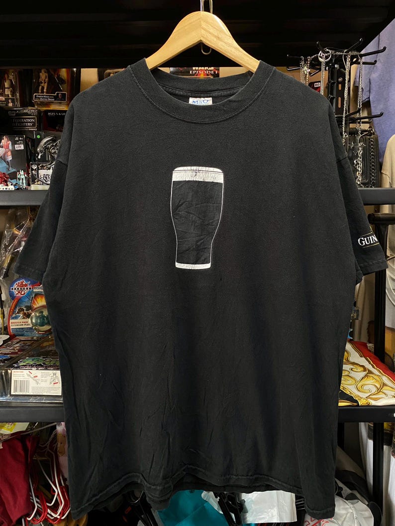 May include: Black t-shirt featuring a white outline of a pint of stout. The shirt has a crew neck and short sleeves. The word "GUIN" is printed on the right sleeve. The shirt is hanging on a wooden hanger.