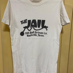 May include: White t-shirt featuring a black graphic that reads "THE JAIL" with a chain and ball design. The address "1105 Bell Grimes Ln. Nashville, Tenn." is printed below the graphic.