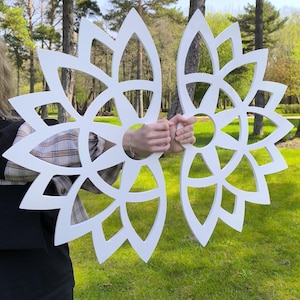 May include: Two white, laser-cut wooden wall decorations in a floral design. The decorations are held up in front of a green grassy background with trees in the distance. The floral design has a circular shape with pointed petals.