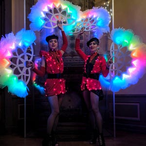 May include: Two performers in red and black costumes hold large, white feathered fans with colorful LED lights. The fans are circular and have a geometric design.