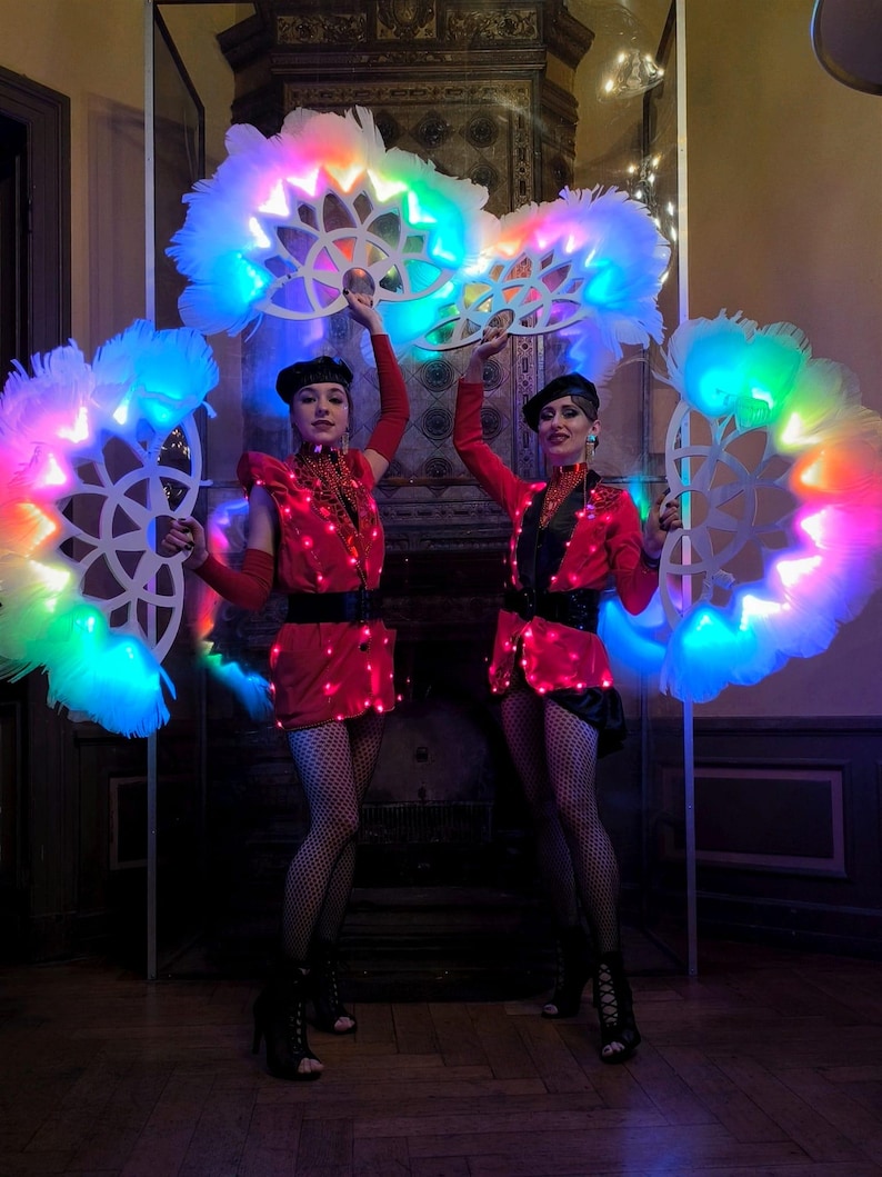 May include: Two performers in red and black costumes hold large, white feathered fans with colorful LED lights. The fans are circular and have a geometric design.