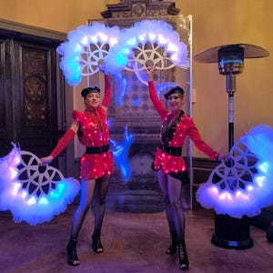 May include: Two performers in red and black costumes with white feathered fans that light up with blue and white lights. The performers are in a room with a fireplace and a heater in the background.
