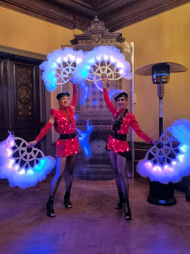 May include: Two performers in red and black costumes with white feathered fans that light up with blue and white lights. The performers are in a room with a fireplace and a heater in the background.