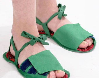 May include: A pair of green sandals with red and blue accents. The sandals have a strap that goes around the ankle and a toe loop. The soles of the sandals are covered in small, red bumps.