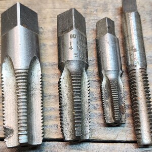 May include: Close-up of five metal tap and die sets. The tap and die sets are silver in color and have a hexagonal shape. The tap and die sets are marked with the text "BUTTER  1/4  NPT  56".
