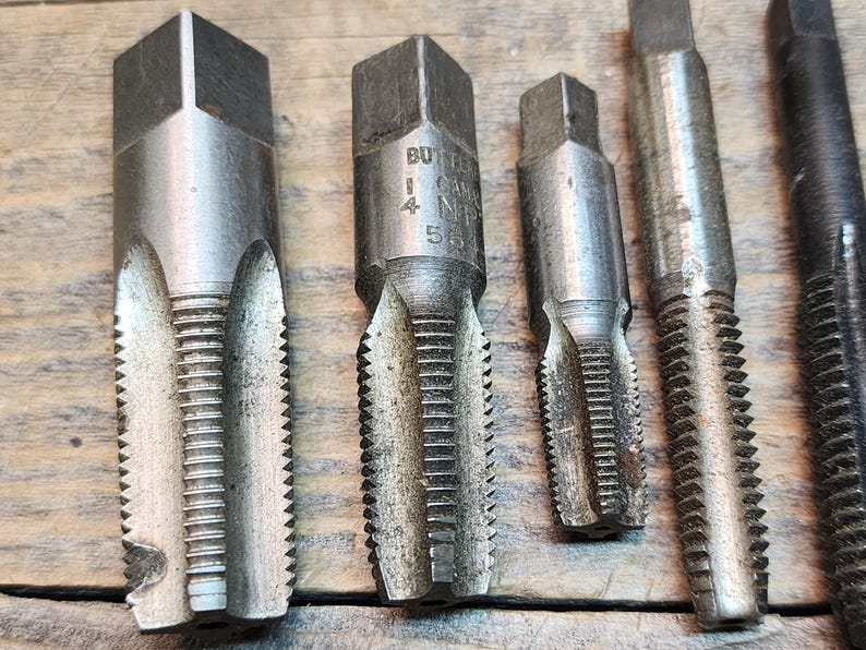 May include: Close-up of five metal tap and die sets. The tap and die sets are silver in color and have a hexagonal shape. The tap and die sets are marked with the text "BUTTER  1/4  NPT  56".