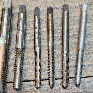 May include: Close-up of seven silver metal tap and die sets with different sizes and thread counts. The tap and die sets are arranged horizontally on a wooden surface.