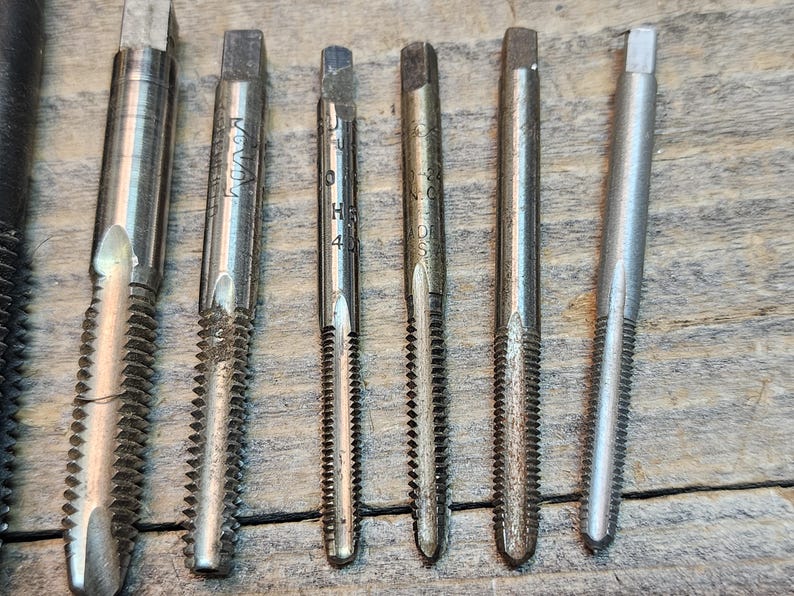 May include: Close-up of seven silver metal tap and die sets with different sizes and thread counts. The tap and die sets are arranged horizontally on a wooden surface.