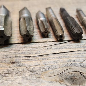 May include: A set of eight metal tap and die sets, used for threading holes and creating nuts. The tools are arranged in a row on a wooden surface.