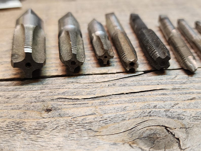 May include: A set of eight metal tap and die sets, used for threading holes and creating nuts. The tools are arranged in a row on a wooden surface.