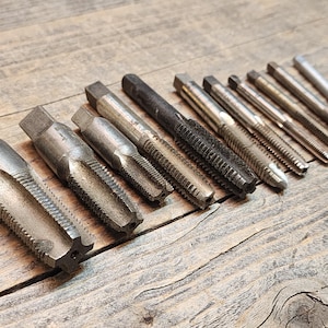 May include: A set of 12 silver-toned metal tap and die tools for threading metal. The tools are arranged in a row on a wooden surface.