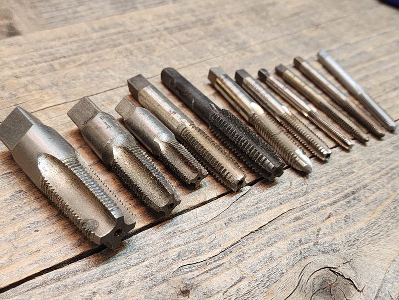 May include: A set of 12 silver-toned metal tap and die tools for threading metal. The tools are arranged in a row on a wooden surface.