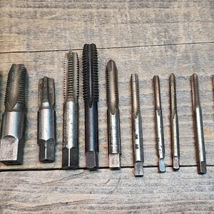 May include: A set of 12 silver and black tap and die tools. The tools are arranged in a row on a wooden surface.