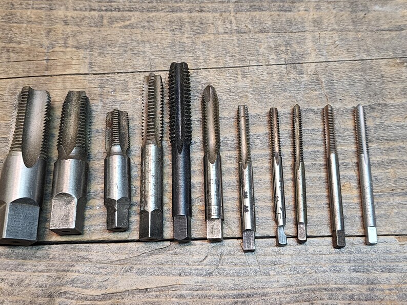 May include: A set of 12 silver and black tap and die tools. The tools are arranged in a row on a wooden surface.