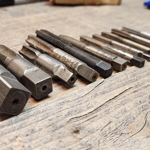 May include: A set of 14 silver-toned metal tap and die sets. The tap and die sets are arranged in a row on a wooden surface.