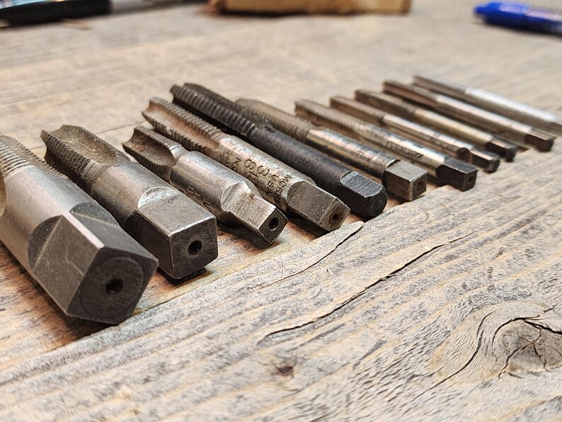 May include: A set of 14 silver-toned metal tap and die sets. The tap and die sets are arranged in a row on a wooden surface.