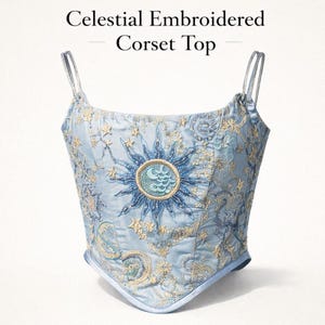 May include: Light blue corset top with celestial embroidery. The top features a sun and moon design with stars and swirls. The text "Celestial Embroidered Corset Top" is at the top of the image.