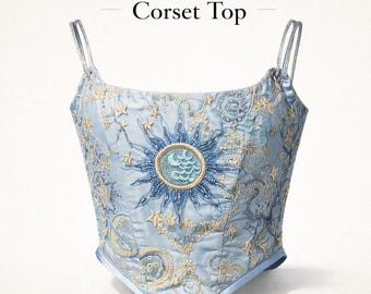 Pode incluir: Top de corset azul claro com bordado celestial. O design apresenta um sol e uma lua, estrelas e redemoinhos. O texto "Celestial Embroidered Corset Top" está no topo da imagem.