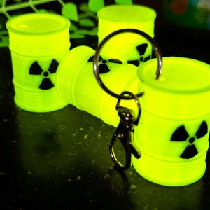 May include: A keychain featuring four bright yellow glow-in-the-dark barrels with black radiation symbols. The barrels are attached to a silver key ring.