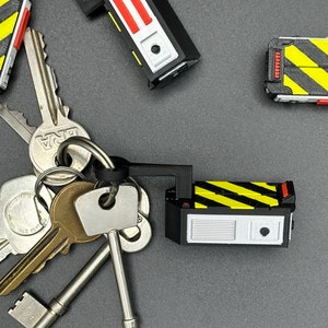 May include: A set of five keychains featuring a black and yellow striped design. The keychains are shaped like small rectangular boxes with a red accent. The keychains are attached to a set of keys.