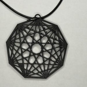 May include: A black geometric pendant necklace. The pendant has a complex, openwork design with a central floral-like pattern. The necklace is suspended from a black cord. The pendant is made of a dark material, possibly metal or plastic.