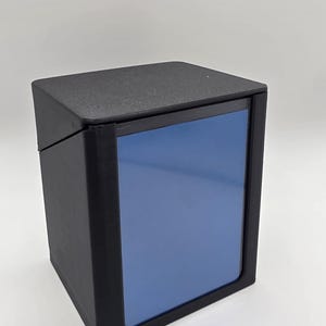 May include: A black, rectangular box with a blue panel on the front. The box has a matte finish and a hinged lid. The design is simple and modern, suitable for storage or display.