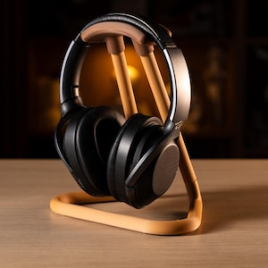 Puede incluir: Auriculares negros de diadema sobre un soporte de madera marrón claro. Los auriculares tienen grandes almohadillas acolchadas y una banda metálica. El soporte tiene una base curva y tres soportes verticales, todos en color marrón claro.