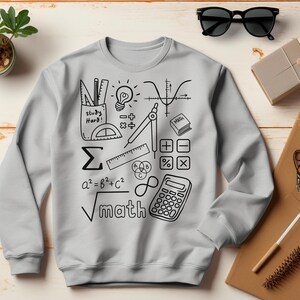 Könnte beinhalten: Hellgraues Sweatshirt mit schwarzem Mathe-Motiv. Die Grafik enthält mathematische Symbole, einen Taschenrechner und die Wörter "study hard" und "math". Eine schwarze Sonnenbrille und eine kleine Box sind sichtbar.