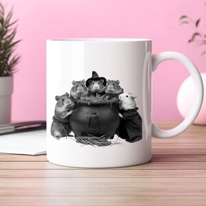 Könnte beinhalten: Weiße Keramiktasse mit einer Schwarz-Weiß-Illustration von Meerschweinchen um einen Kessel. Ein Meerschweinchen trägt einen Hexenhut, ein anderes einen schwarzen Umhang. Die Tasse hat einen gebogenen Henkel.