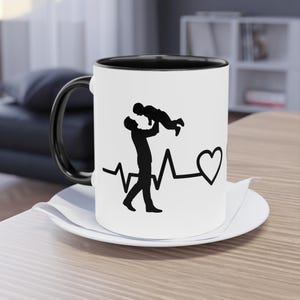 Op de afbeelding: Witte keramische koffiemok met een zwarte binnenkant en handvat. De mok heeft een zwart silhouet van een ouder die een kind vasthoudt, met een hartslaglijn en een hartsymbool. De mok staat op een wit schoteltje.