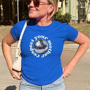 Puede incluir: Una camiseta de manga corta en color azul real con un gr&aacute;fico central de una bola de discoteca plateada rodeada por el texto circular blanco "respect your mother". La prenda se combina con vaqueros azul claro y un bolso peque&ntilde;o color crema.
