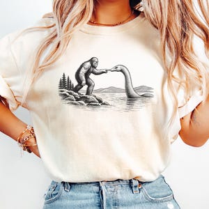 May include: A cream-colored short-sleeve t-shirt featuring a black ink-style graphic of Bigfoot standing on a rocky shore and touching fingers with the Loch Ness Monster in a lake, with pine trees and distant mountains in the background.