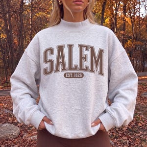 May include: A light gray heathered sweatshirt with a mock neck and long sleeves. The front features the text SALEM in large brown block letters with a white outline, and the text EST. 1626 inside a brown oval shape below the main lettering.