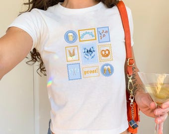 Puede incluir: Top corto blanco con un gráfico colorido con temática de Oktoberfest. El diseño incluye jarras de cerveza, pretzels, notas musicales y la palabra "proost!" en un estilo de sello postal. La camiseta es de manga corta y tiene cuello redondo.