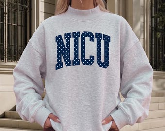 Puede incluir: Una sudadera gris claro jaspeado con cuello alto y mangas largas. El frente presenta el texto NICU en grandes letras de bloque azul oscuro con un patrón de puntos blancos. La sudadera tiene un ajuste relajado con puños y dobladillo acanalados.