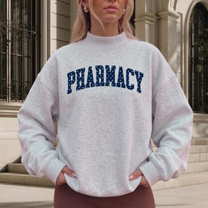 May include: A light gray heathered sweatshirt with a mock neck and long sleeves. The word PHARMACY is printed across the chest in a dark navy blue collegiate font, featuring a pattern of small white dots within the letters.